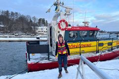 En man i sjöräddningsuniform står framför en räddningsbåt vid en snötäckt hamn.