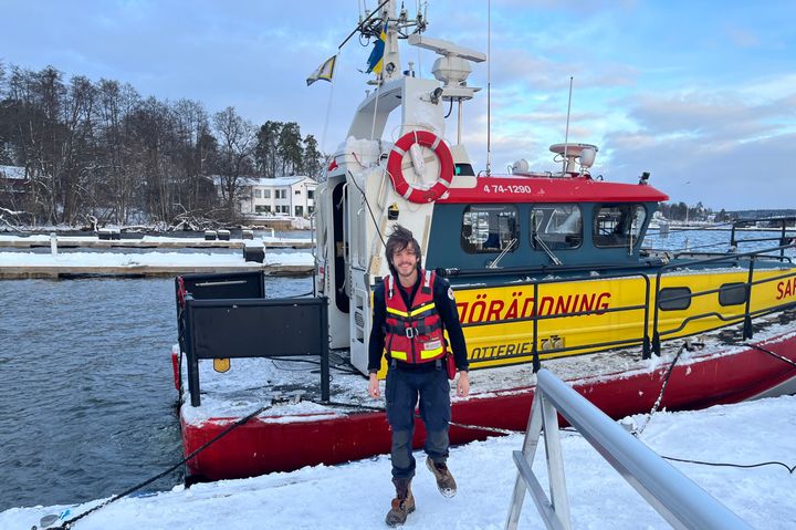 En man i sjöräddningsuniform står framför en räddningsbåt vid en snötäckt hamn.