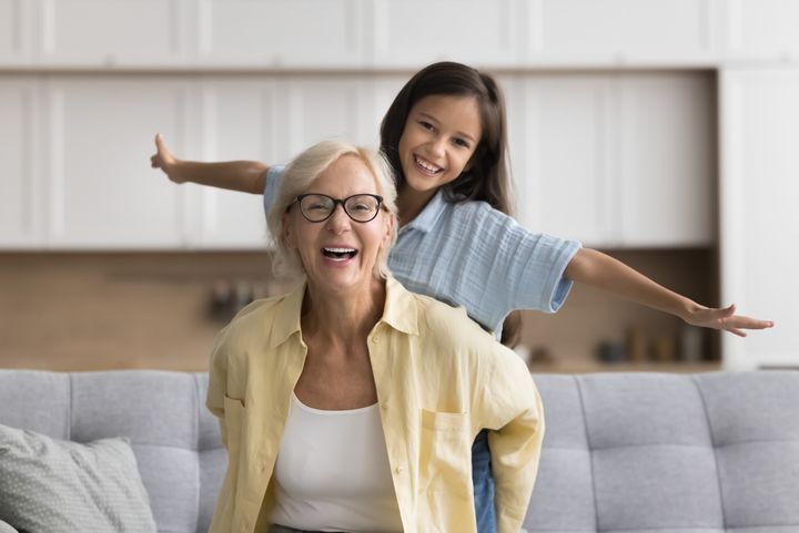 En äldre kvinna och en ung flicka skrattar och gör flygplansposer i ett vardagsrum.