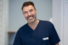 Person i sjukvårdsuniform ler framför kameran i en kontorsmiljö.