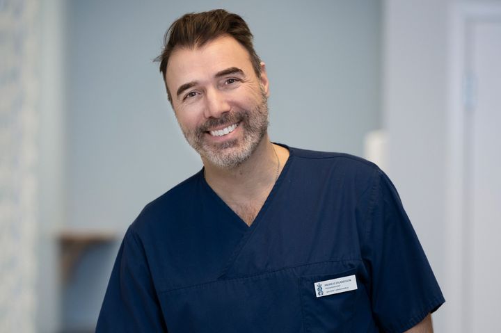 Person i sjukvårdsuniform ler framför kameran i en kontorsmiljö.