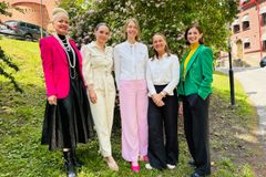 En grupp med fem leende kvinnor står utomhus framför grönska och en tegelbyggnad. De är klädda i färgglada och eleganta kläder.