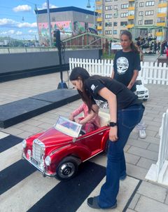 Två ungdomar i svarta t-shirts med texten Tigrarna hjälper ett barn i röd leksaksbil.