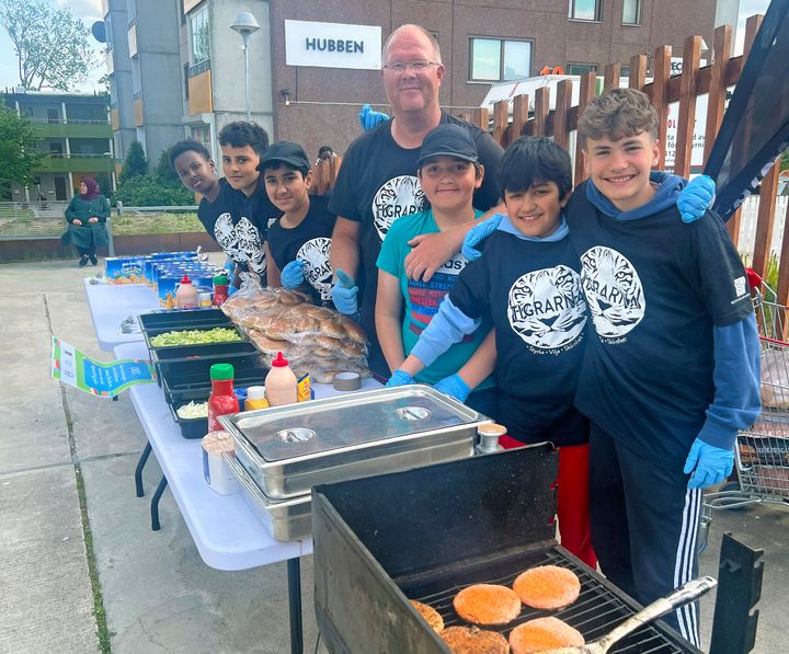 Sex ungdomar och en vuxen i svarta t-shirts med texten Tigrarna grillar hamburgare med tillbehör.