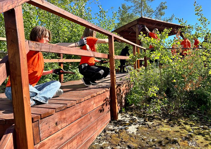 En grupp ungdomar i orange t-shirt oljar in en träkonstruktion utomhus på sommaren.
