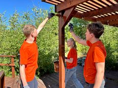 En grupp ungdomar i orange s-hirts oljar in taket på en träkonstruktion utomhus.