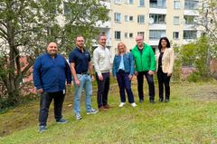 Sex personer står i en skogsdunge framför ett långt hyreshus.