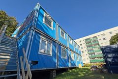 Blå kontorsbodar framför flerbostadshus i vitt och grönt.