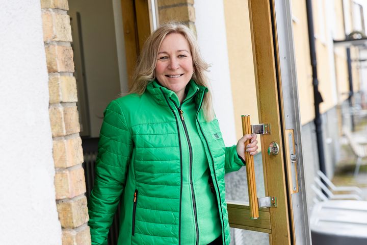 En leende kvinna i grön jacka går ut genom en entréport.