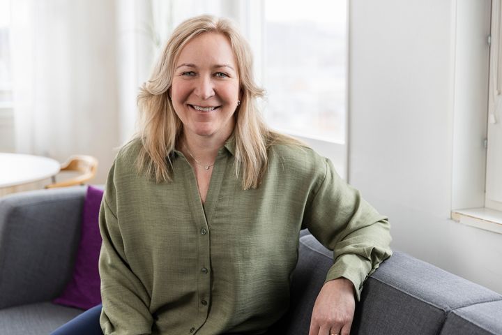 En blond kvinna i grön skjorta sitter i en grå soffa framför ett fönster.