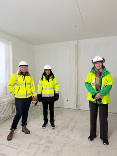 Tre kvinnor med vita bygghjälmar, två med gul varseljacka och en med gul varselväst inne på ett bygge.