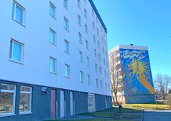 Dela av vitt flervåningshus samt ett hus i bakgrunden med färgglad fasadmålning på gaveln.