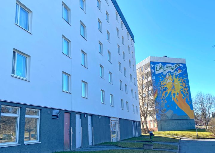 Dela av vitt flervåningshus samt ett hus i bakgrunden med färgglad fasadmålning på gaveln.