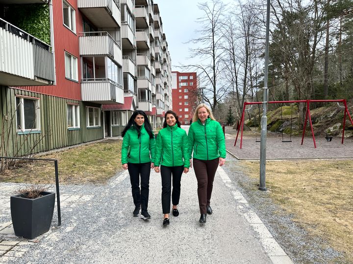 Tre kvinnor i gröna jackor går på en gångväg framför ett rött höghus med grön sockel.