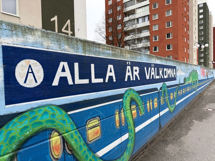 Målad mur där en orm slingrar sig runt en tunnelbanevagn. I bakgrunden syns färgglada höghus.