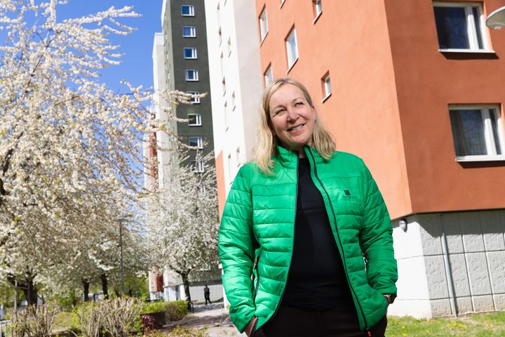 Kvinna i grön jacka framför höghus i olika färder, blommande träd bredvid.