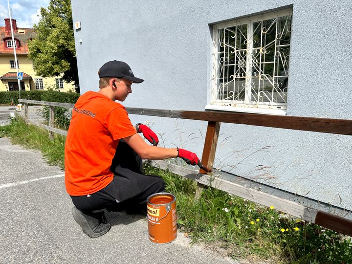 En ung person i orange t-shirt och svart keps sitter på huk och målar ett staket vid en parkeringsplats