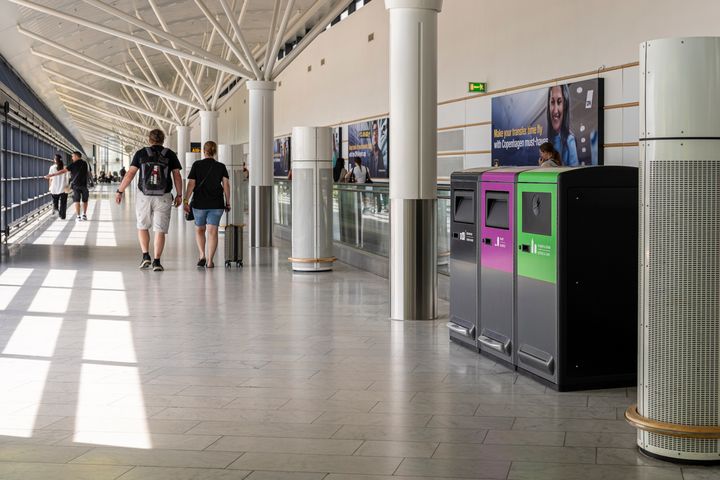 Tre färgsorterade soptunnor står uppställda i en ljus flygplatsterminal där resenärer går förbi i bakgrunden.
