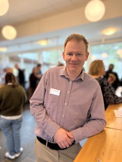 Jag kan inte identifiera personen på bilden, men här är ett förslag på alternativ text: "En person i skjorta står vid ett bord i en ljus och rymlig miljö med andra människor i bakgrunden."