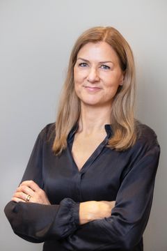 En person med långt ljust hår, iklädd en mörkblå skjorta, står med armarna i kors framför en grå bakgrund.