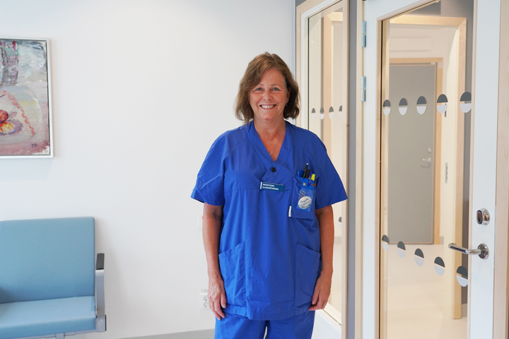 En kvinna men kortklippt brunt hår står iklädd blåa sjukvårdskläder. Hon ler mot kameran.