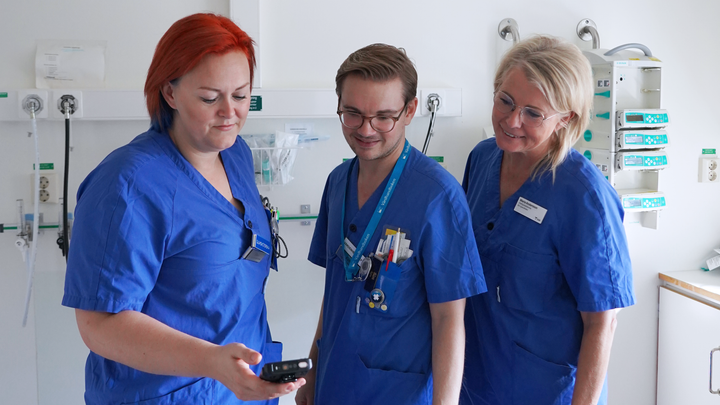 Tre personer i sjukvårdskläder står bredvid varandra. En av dem håller i en mobiltelefon. Alla kollar i mobiltelefonen.
