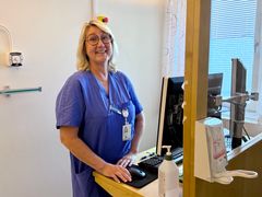 Sjuksköterska står framför en dator och ler mot kameran.