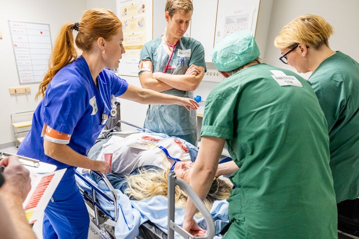 Tre vårdmedarbetare i gröna kläder, en vårdmedarbetare i blå kläder tar hand om skadad patient som ligger på en brits i sjukhusmiljö.