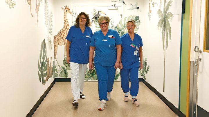 Tre vårdpersonal i blå arbetskläder går tillsammans i en korridor med väggar dekorerade med djungelmönster och djurillustrationer.