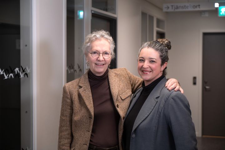 Två kvinnor står bredvid varandra i ett kontorsutrymme, båda leende och klädda i kavaj.