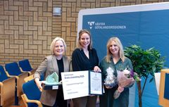 Tre personer poserar med pris och blommor framför en skylt från Västra Götalandsregionen.