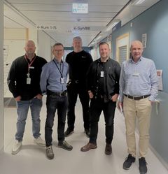 Fem personer står i en sjukhuskorridor med skyltar för rum och avdelningar i bakgrunden.