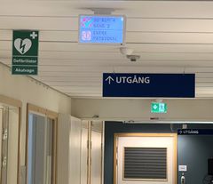 Informationspanel i taket på sjukhus som visar vårdkallelse, med skyltar för utgång och defibrillator bredvid.