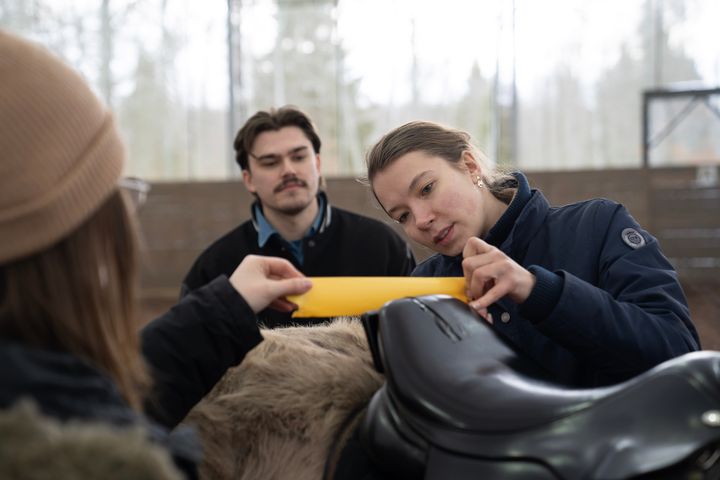 Tv%E5%20personer%20placerar%20en%20gul%20niv%E5m%E4tare%20p%E5%20en%20sadel%20medan%20en%20tredje%20person%20tittar%20p%E5.