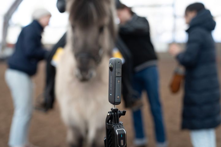 En%20kamera%20monterad%20p%E5%20ett%20stativ%20med%20en%20suddig%20h%E4st%20och%20tre%20m%E4nniskor%20i%20bakgrunden.