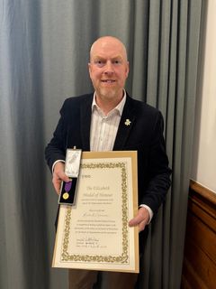 En man i kavaj håller en medalj och ett diplom som lyder "The Elizabeth Medal of Honour 2026".