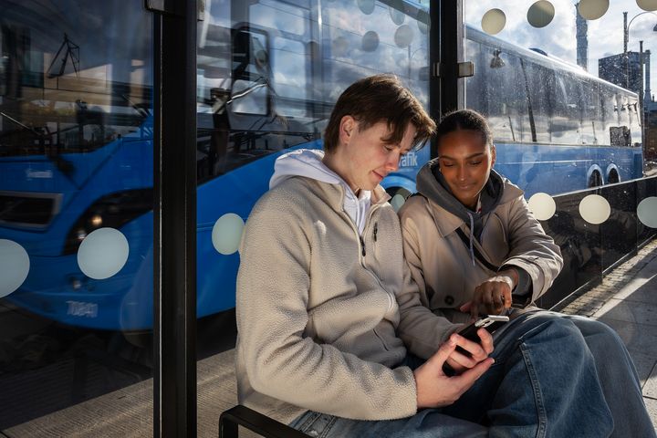 Två personer sitter på en busskur vid en hållplats. En blå buss står parkerad bakom glasväggen. Personerna sitter bredvid varandra och tittar på en mobiltelefon som en av dem håller i, medan den andra pekar på skärmen.