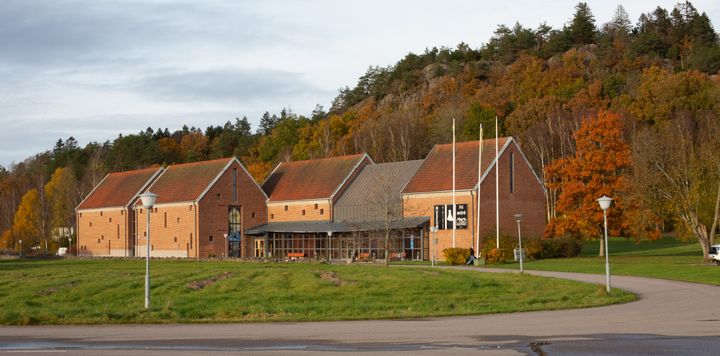 Byggnadskomplex i tegel med sluttande tak omgiven av gräsyta och träd, Lödöse museum.