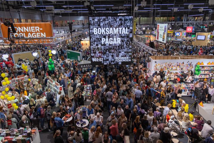 En stor folkmassa på en bokmässa med många montrar och ett stort skylt där det står "Boksamtal pågår".
