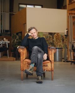 Person sitter i sliten brun fåtölj i ateljé med benen korsade. Tavlor och arbetsytor i bakgrunden.