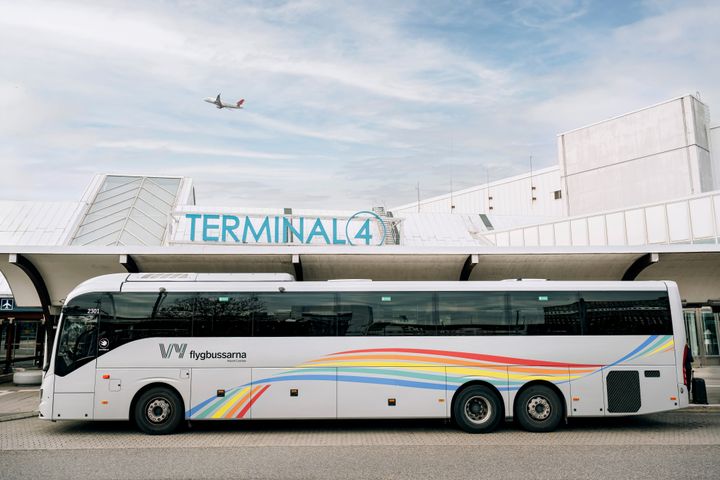 En buss från Vy flygbussarna står framför Terminal 4 på en flygplats, ett flygplan syns i luften ovanför.