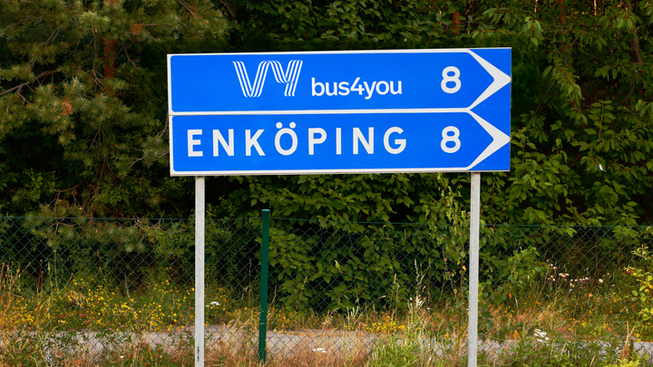 Vägskylt med texten "Enköping 8" och "bus4you 8". Bakgrund av träd och buskar.