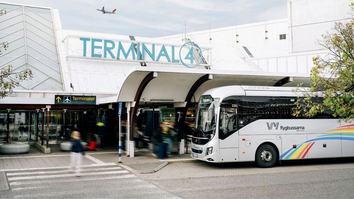 Buss från Vy flygbussarna står utanför Terminal 4 vid en flygplats. Ett flygplan syns i luften.