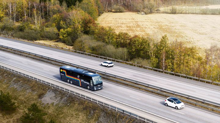 En långfärdsbuss kör på en landsväg med två personbilar. Skog och åkrar omger vägen.