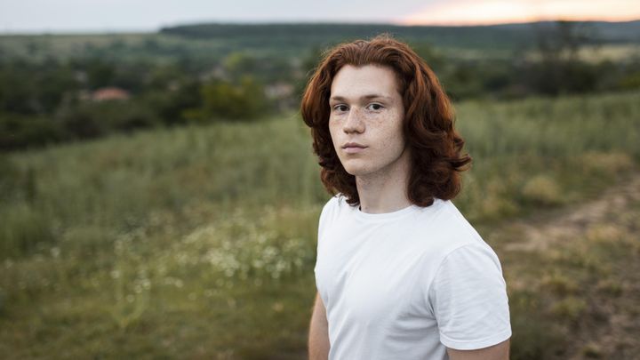 Ung man med långt rött hår står på en fält med landsbygd i bakgrunden.