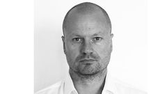 Bilden visar en person med kortklippt hår och en skäggstubb, som tittar in i kameran mot en vit bakgrund.