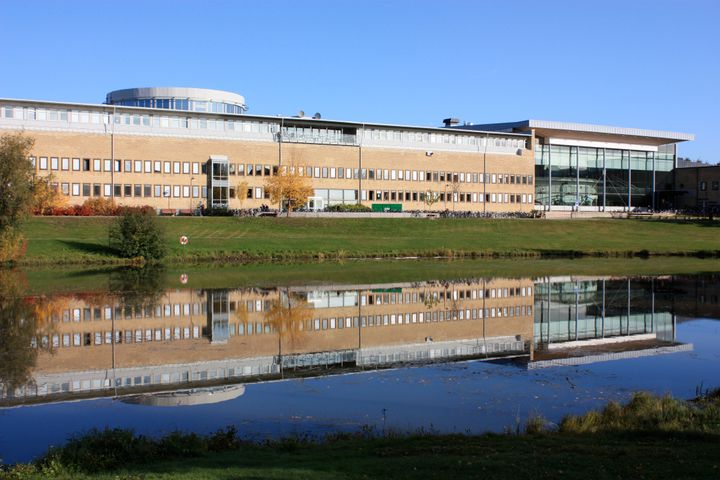 Universitetsbyggnad med glasfasad speglas i en stilla damm under en klarblå himmel.