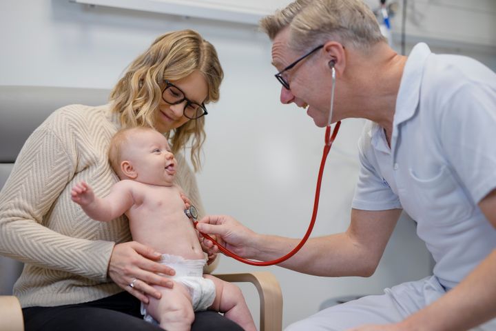 En läkare undersöker en bebis som sitter i sin mammas knä.