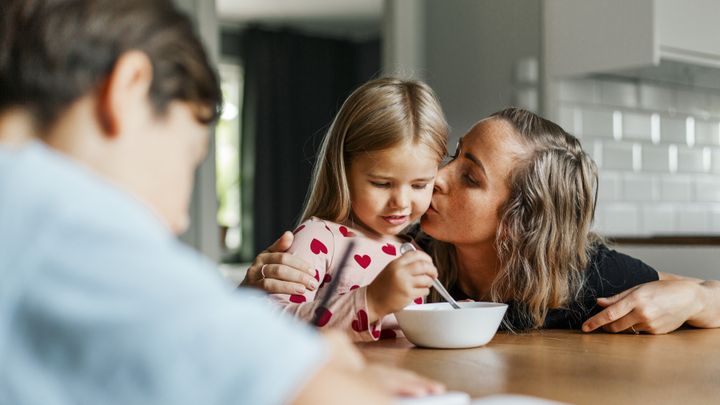 Mor kysser sin dotter som äter frukost vid ett köksbord; en man syns i förgrunden.