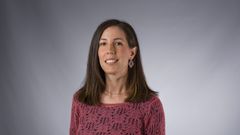 En person med långt, mörkt hår står mot en grå bakgrund och bär en rosa tröja.